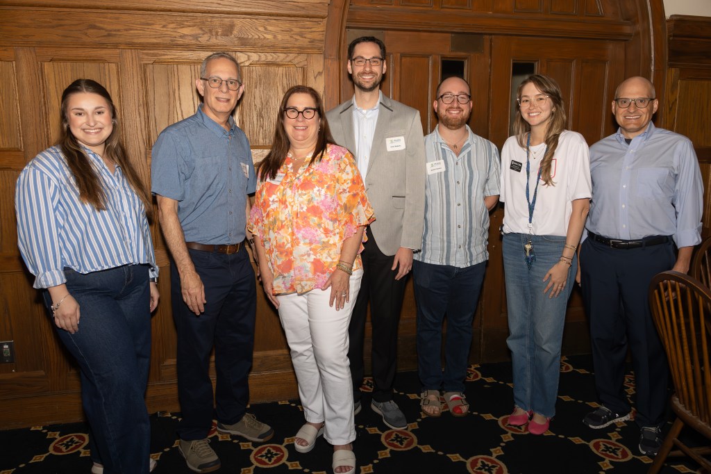 A group of seven people posing together in a stylish indoor setting, showcasing a warm, welcoming atmosphere. They are all smiling and dressed in casual attire, with a wooden wall and patterned carpet in the background.