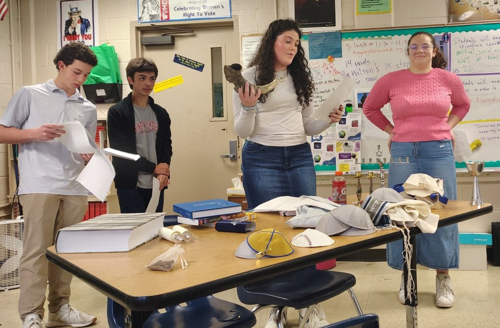 A diverse group of four students stands in a classroom, participating in a presentation. The student in the center holds a shofar and a paper, while others hold notes and various items are displayed on a table, including books and religious items.