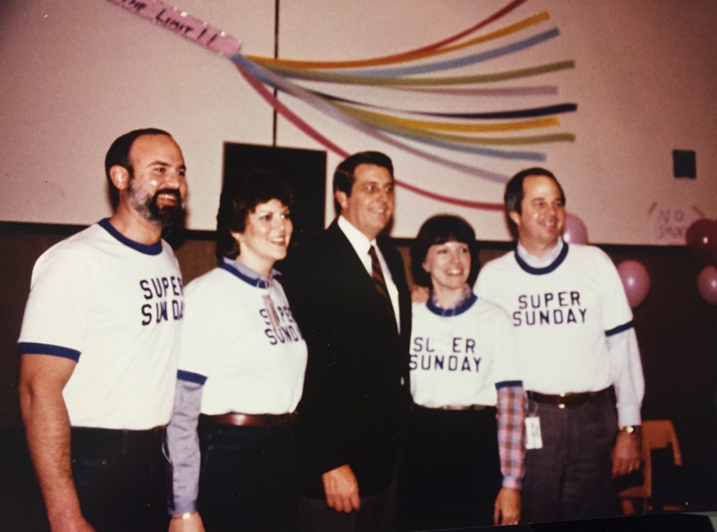 A group of four people posing for a photo wearing 'Super Sunday' t-shirts, smiling and standing in a festive setting decorated with colorful streamers.