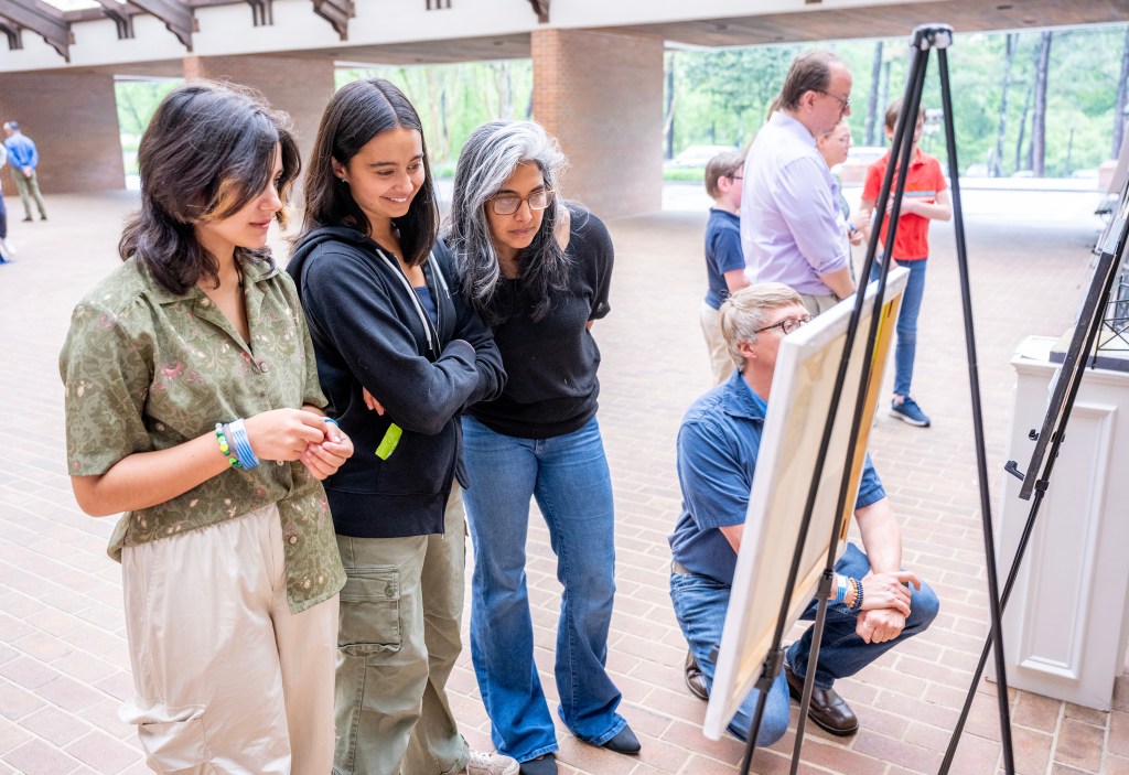 A group of individuals, including three young women, are gathered in an indoor setting looking at artwork displayed on easels. Two women are standing closely together, observing the piece, while a third is pointing at it. In the background, several other people engage with different artworks.