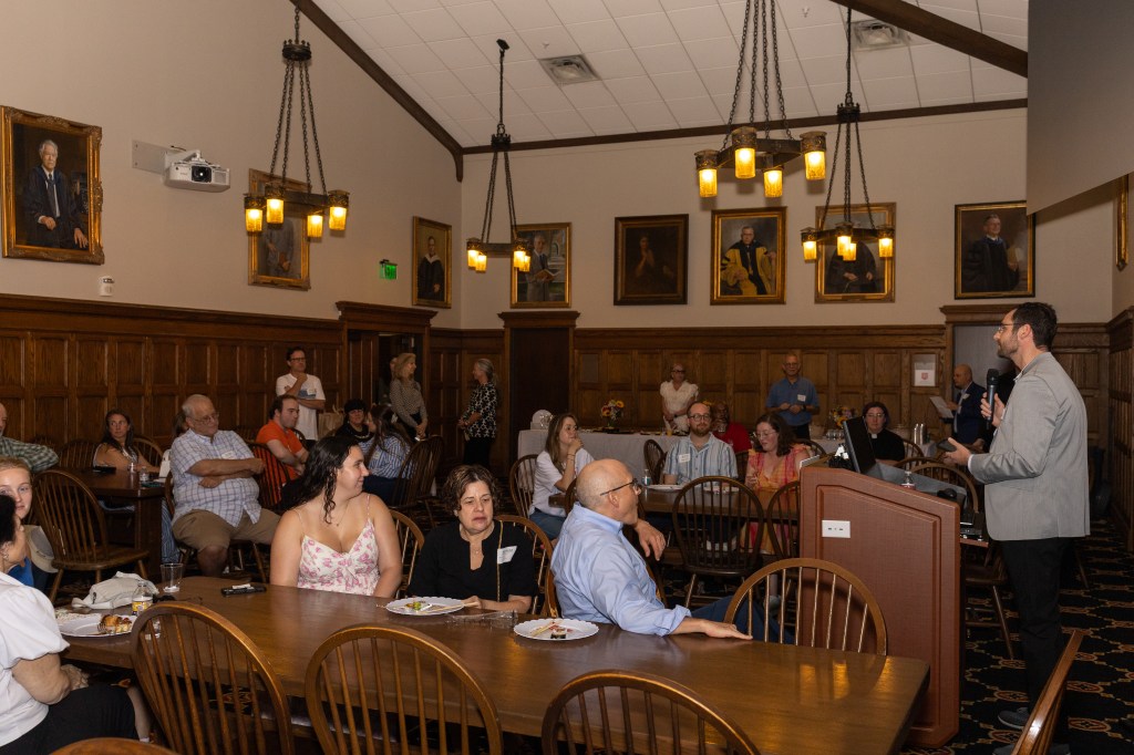 A warm reception event for incoming Jewish students at Rhodes College, featuring attendees seated at wooden tables, with a speaker addressing the audience in a historic hall adorned with portraits.