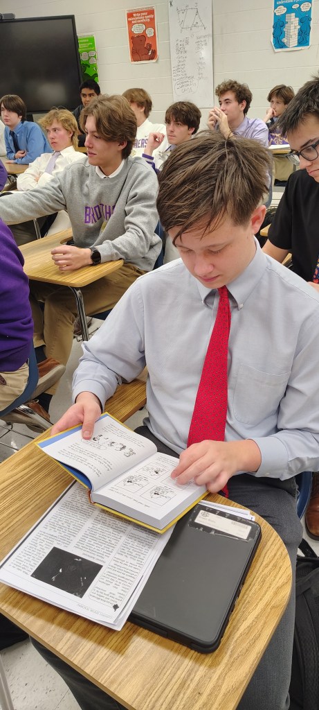 A high school student in a dress shirt and tie reads a book at a desk, while other students are engaged in the background, during a class session.
