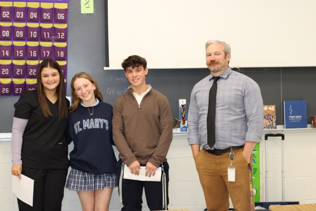 Four students standing in a classroom setting, with educational materials and a chalkboard in the background.