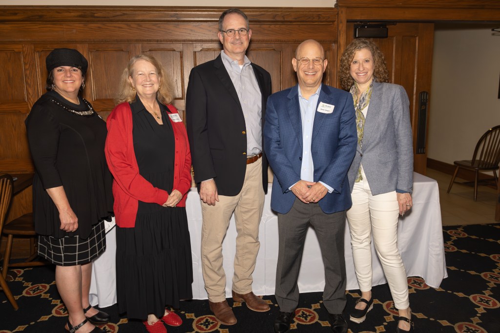 Five individuals posing together for a photo at an event, with a wooden backdrop and tables visible in the background.