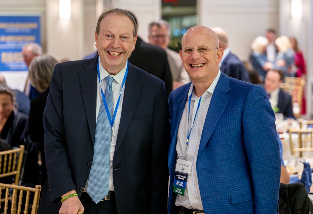 Two smiling men pose for a photo together at a fundraising event, wearing formal attire and name tags, with other attendees visible in the background.