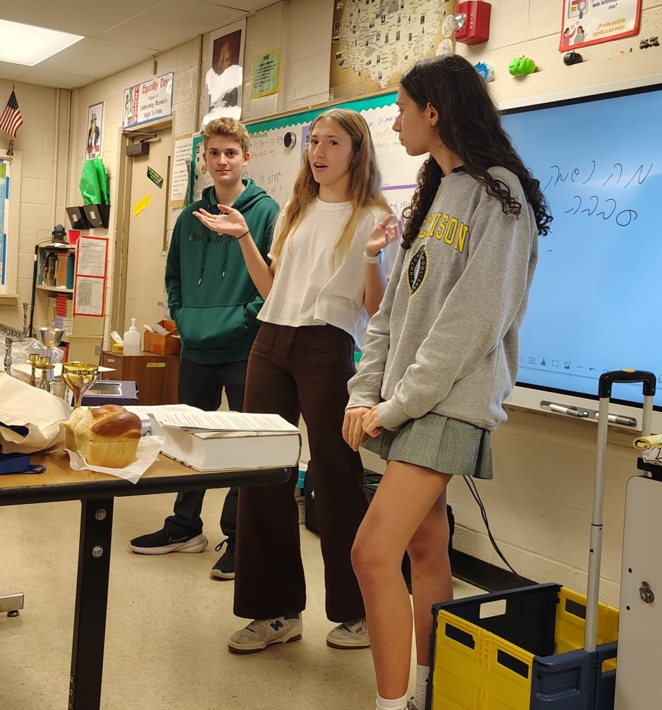 Three students presenting in a classroom, with one girl speaking while the others listen. On the table are various items including a loaf of bread and books. A whiteboard is visible in the background.